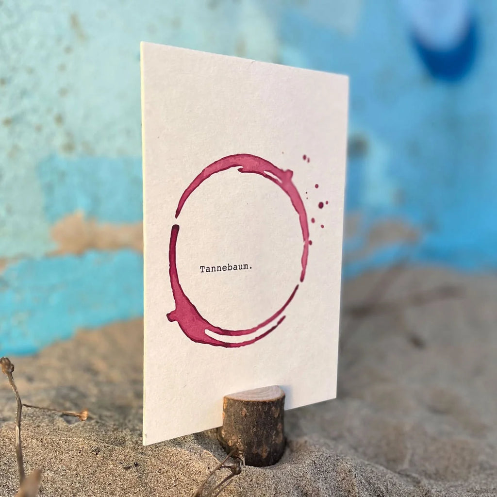 Postkarte mit Rotweinfleck-Ring und Wort Tannebaum im Sand vor blauer Wand, witzige Weihnachtskarte | Prinzenart